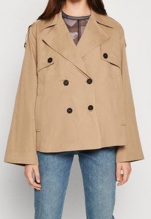 Femme portant une veste beige croisée à boutons noirs et un jean bleu, debout devant un fond uni.