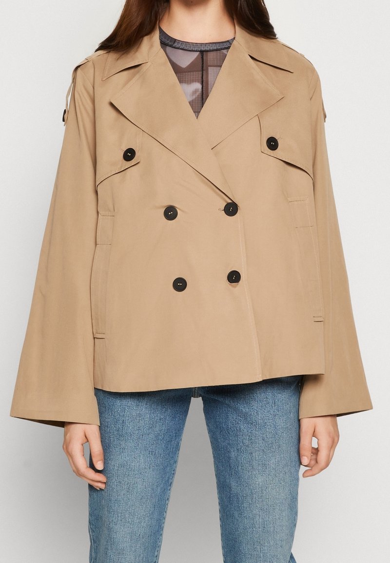 Femme portant une veste beige croisée à boutons noirs et un jean bleu, debout devant un fond uni.