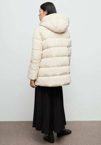 Beige pufferjack met een capuchon, met gequiltte secties en een relaxed fit. Draagt over een lange zwarte rok, met simpele zwarte schoenen.