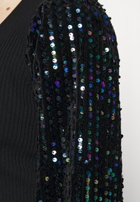 Haut noir avec un corps côtelé texturé et des manches bouffantes ornées de sequins, présentant des sequins irisés aux teintes vertes, bleues et argentées.