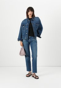 Veste en denim avec de grandes poches avant, de couleur bleu foncé, portée sur un haut noir ; assortie à un jean droit bleu clair et des sandales noires. Tenant un sac violet clair.