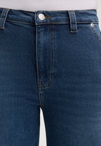 Jeans in denim blu scuro con un classico design a cinque tasche, cuciture visibili e una chiusura con bottone metallico in vita.