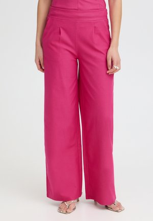 Woman wearing high-waist wide-leg pink pants and open-toe heels with rings on left hand, standing against a plain light background.