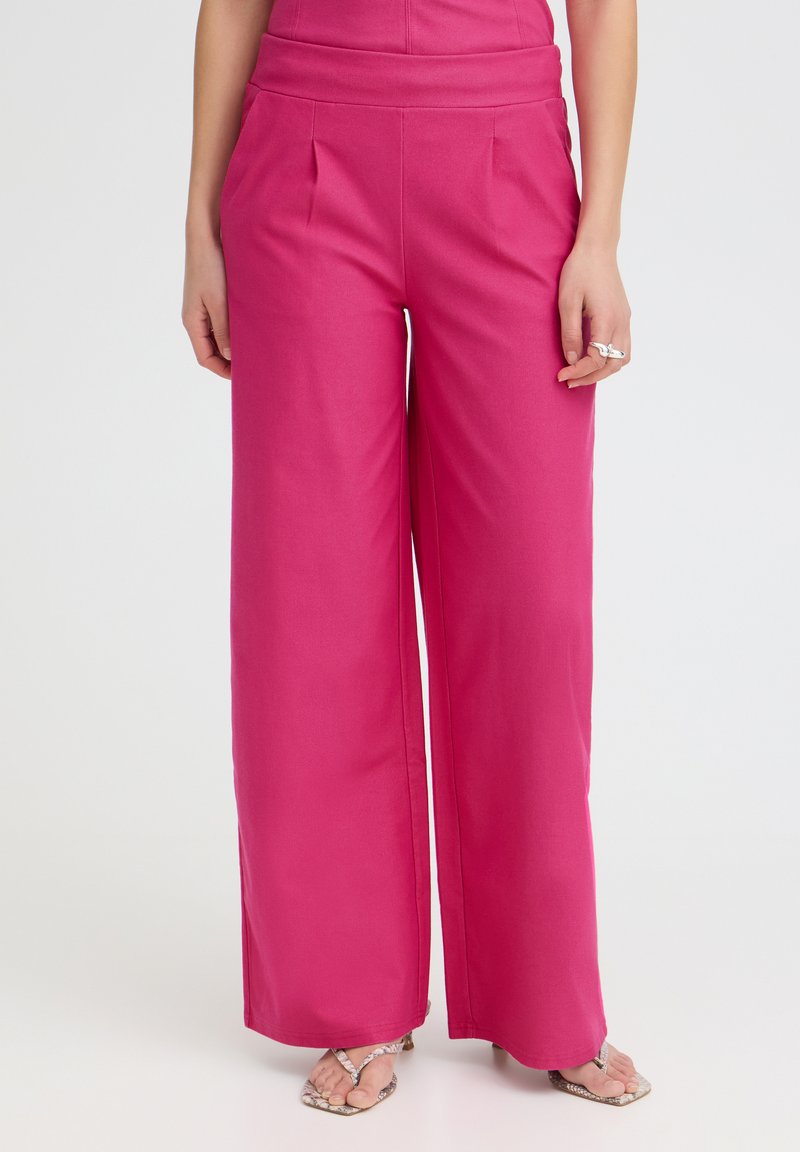 Femme portant un pantalon rose taille haute à jambes larges et des talons ouverts avec des bagues à la main gauche, debout devant un fond clair uni.