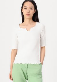 White ribbed top with a scalloped hem and sleeves, featuring a rounded neckline with a keyhole detail. Paired with green pants.