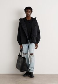 Black hooded jacket, black t-shirt, ripped light blue jeans, and black shoes. Holding a black tote bag with "HUGO" printed on it.