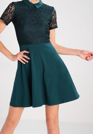 Vrouw draagt een knielange donkerteal jurk met korte kanten mouwen en een kraag, poseert met één hand op de heup en een arm uitgestrekt.