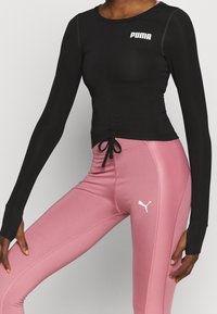 Frau in einem schwarzen langen Puma-Oberteil und hochtaillierten rosa Puma-Leggings, die mit einem gebeugten Bein steht und die Hände an den Hüften hat.