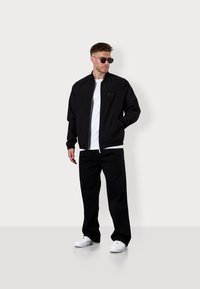 Schwarze Bomberjacke mit Reißverschluss und Seitentaschen, getragen über einem weißen Hemd. Kombiniert mit schwarzen Hosen und weißen Sneakern.