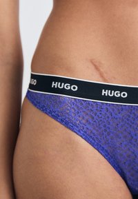 Paarse kanten string met zwarte elastische tailleband en witte "HUGO" logo. Close-up toont de textuur van de stof en details.