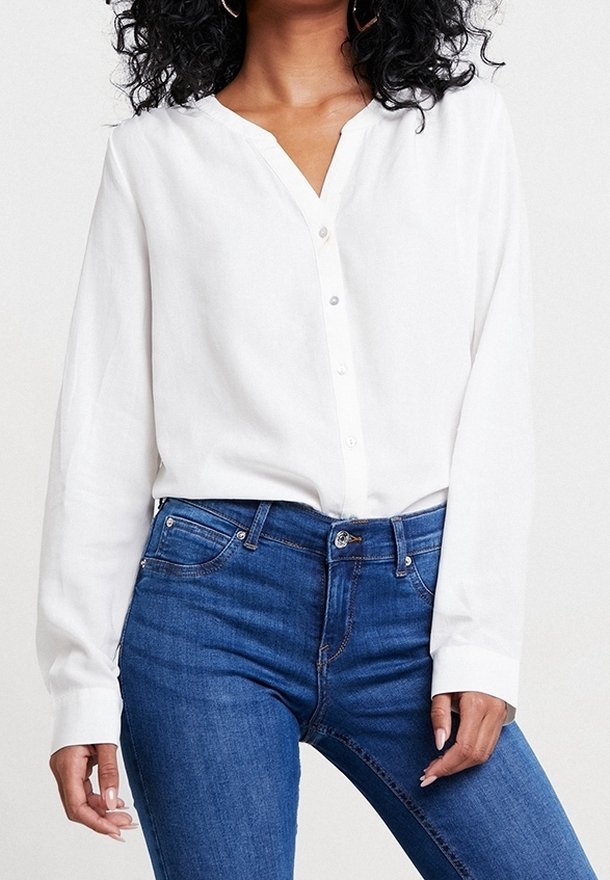 Femme portant un chemisier blanc boutonné à manches longues et un jean bleu, posant une main le long du corps devant un fond uni.