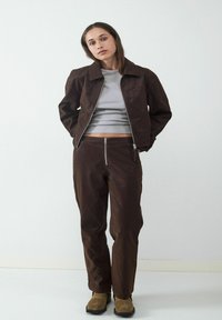 Ciemnobrązowa kurtka z kołnierzykiem, połączona z pasującymi spodniami z kieszeniami na zamek. Model nosi szary top z krótkim rękawem.