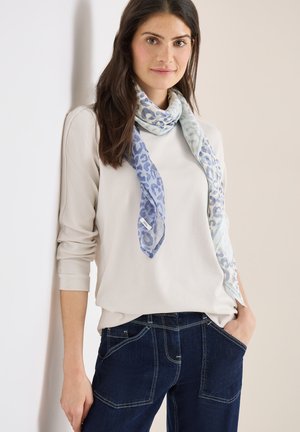 Ung kvinde med langt mørkt hår iført en hvid top, blå leopardmønstret tørklæde og mørkeblå jeans, stående med den ene hånd i lommen.