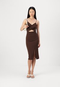 Hnedé midi šaty s výrezom, tenkými pásikmi a predným rozparkom. Modelka nesie malú bielu kabelku a má na sebe béžové sandále.