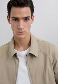 Chaqueta ligera beige con cuello, que presenta un sutil logo de calavera. Llevada sobre una camiseta blanca, con una tela suave y un diseño moderno.