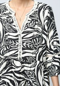 Blouse à motifs noir et blanc avec un col en V, une patte de boutonnage décorative et des manches bouffantes. Présente des rayures verticales et des motifs de feuilles.