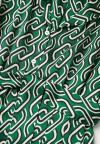 Tissu de chemise verte présentant un motif abstrait de lignes entrelacées en noir et blanc, avec des boutons blancs visibles. Texture lisse.