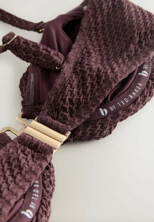 Burgundy textured bra featuring woven fabric with gold accents, adjustable straps, and gold hardware clasp. Brand name visible on the band.