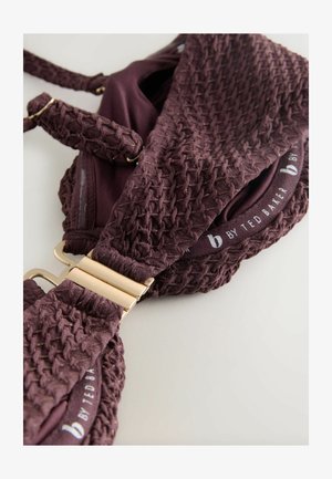 Burgundy textured bra featuring woven fabric with gold accents, adjustable straps, and gold hardware clasp. Brand name visible on the band.