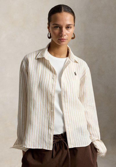 Mujer con el cabello recogido lleva una camisa de botones a rayas beige y blanca sobre una camiseta blanca y pantalones marrones, posando frente a un fondo neutro.