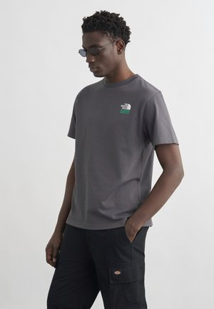 Homme aux cheveux courts et bouclés portant des lunettes de soleil foncées, un t-shirt gris The North Face et un pantalon cargo noir, debout avec une main dans la poche.
