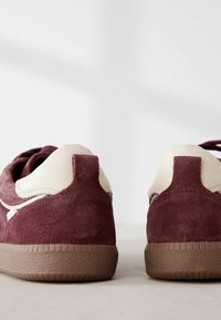 Paire de baskets en daim bordeaux avec des accents en cuir blanc au talon et des semelles en caoutchouc marron, vues de dos sur une surface claire.