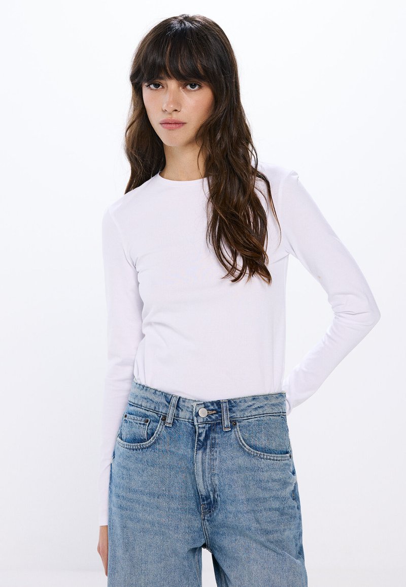 Camicia bianca a maniche lunghe realizzata in tessuto leggero, abbinata a jeans in denim blu a vita alta con tasche anteriori e chiusura a bottone.