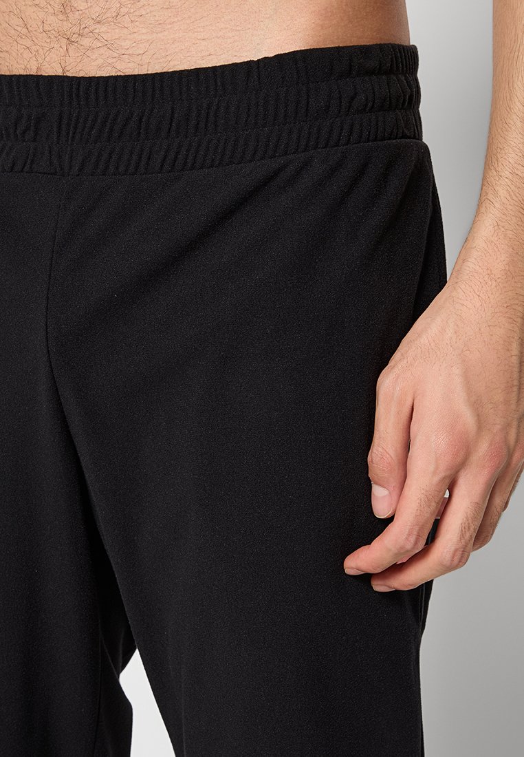 Pantalones deportivos negros con una cintura elástica y una textura de tela suave. La vista en primer plano incluye una mano descansando en la zona del muslo.