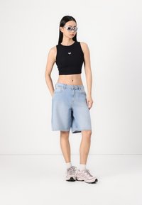 Svart croppet tank top med liten logo, lyseblå denim Bermuda-shorts og rosa joggesko med hvite sokker, plassert mot en nøytral bakgrunn.