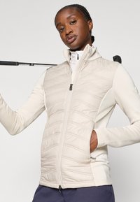 Ljusbeige väst med quiltad framsida, släta sidor, dragkedjestängning, hög krage och sidofickor, bärs med en vit polotröja och en golfklubba.