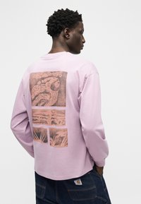 Άνδρας που φοράει ανοιχτό μωβ πουκάμισο με μακριά μανίκια και αφηρημένο κόκκινο-καφέ γραφικό στο πίσω μέρος, συνδυασμένο με σκούρο μπλε τζιν, πόζα μπροστά σε λευκό φόντο.