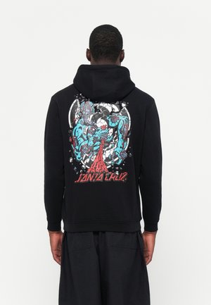 Personne vue de dos portant un sweat à capuche noir avec un graphisme coloré représentant un monstre bleu qui déchire une forme ovale, avec un texte rouge en dessous.