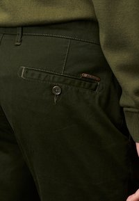 Pantalon en coton vert foncé avec une poche arrière à fermeture par bouton et une petite étiquette en métal. Texture lisse, coupe ajustée.