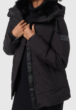 Henkilö, jolla on musta toppatakki, jossa on turkisvuorattu huppu ja vetoketju, pukeutuneena mustan ribbineuleen päälle.