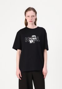 Black cotton t-shirt with loose fit, featuring a graphic print of stylized characters and "KARL" text in contrasting white. Short sleeves.