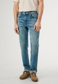 Jeans de mezclilla azul claro con un corte recto, cinco bolsillos y un ligero desgaste. Combinados con zapatillas marrones y una camisa beige.