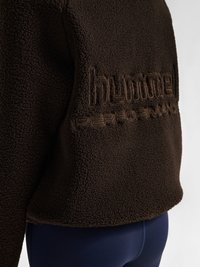 Mørkebrunt tekstureret fleecejakke med hævet broderet 'hummel PERFORMANCE' logo på den nederste ryg, båret med mørkeblå tætsiddende bukser.