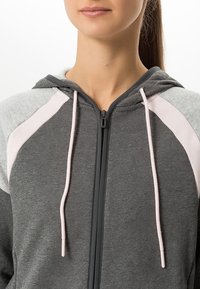 Femme portant un sweat à capuche zippé gris et rose clair avec cordons, vue du cou au haut du torse.