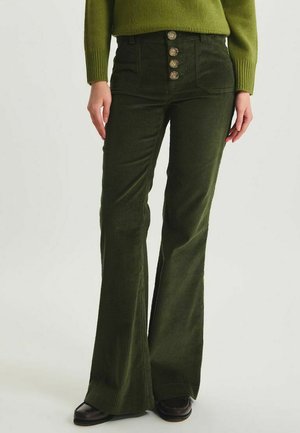 Pantalon en velours côtelé vert foncé taille haute avec jambes évasées et quatre boutons à texture bois, porté avec un pull vert assorti et des chaussures noires.
