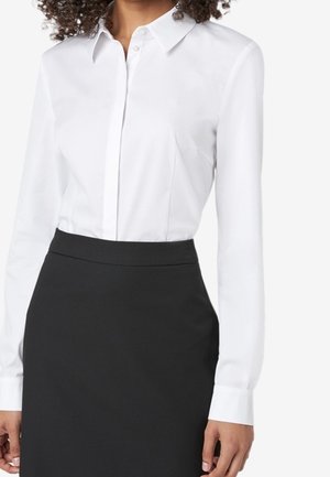 Camicia bianca con colletto e maniche lunghe, abbinata a una gonna a matita nera. La camicia ha una texture liscia e un design aderente.