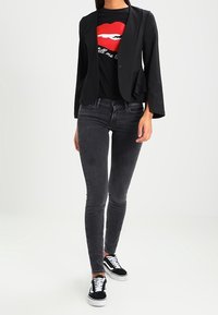 Blazer negro con un diseño ajustado, detalles superpuestos; combinado con una camiseta gráfica negra que presenta labios rojos, y vaqueros skinny gris oscuro, complementados con zapatillas negras.