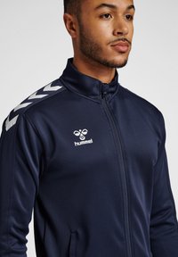 Giacca a track navy con cerniera completa, collo alto e motivi a chevron bianchi sulle spalle. Presenta un logo sul petto e tasche laterali.