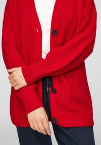 Personne portant un cardigan en maille rouge vif boutonné sur un haut blanc et un pantalon foncé, bras croisés à la taille.