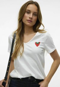 T-shirt en coton blanc avec un décolleté en V et des manches courtes, arborant un design coloré de fraise sur la poitrine gauche. Porté avec un jean sombre et une sangle noire.
