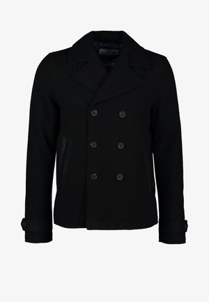 Schwarzer doppelt geknöpfter Peacoat mit breiten Revers, getragen über einem schwarzen Hemd, kombiniert mit dunklen Jeans und schwarzen genieteten Stiefeletten.