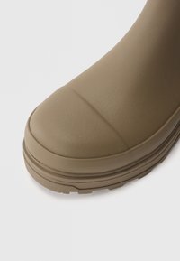 Bota de goma marrón con punta redondeada y superficie texturizada. Presenta una suela acanalada y un acabado suave, ligeramente brillante.