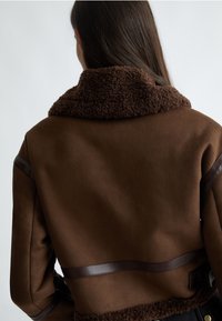 Giacca in shearling marrone con un colletto morbido e testurizzato e dettagli in pelle marrone. Il design presenta una silhouette aderente e linee pulite.