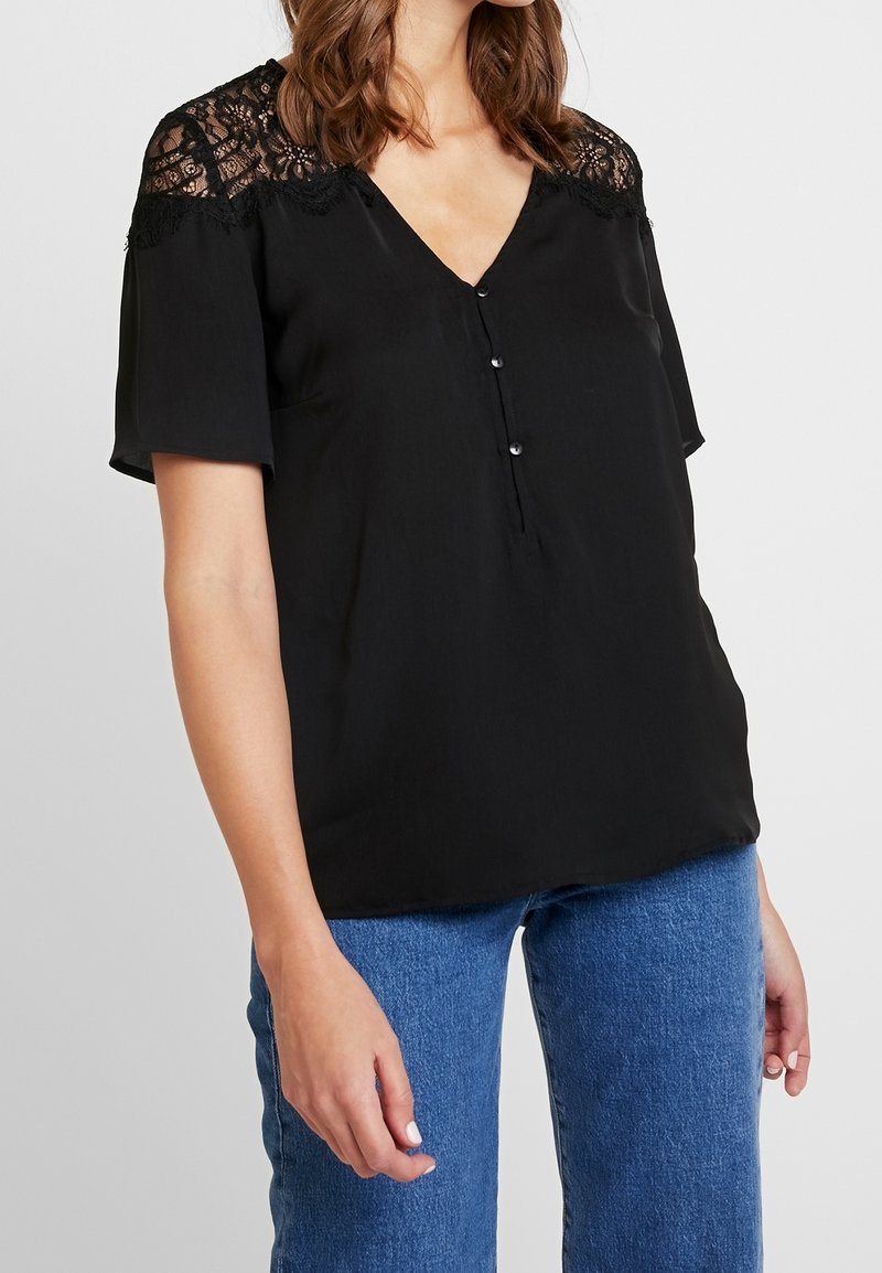 Blusa nera con maniche corte e dettagli in pizzo sulle spalle, caratterizzata da un scollo a V e tre bottoni decorativi sul fronte.
