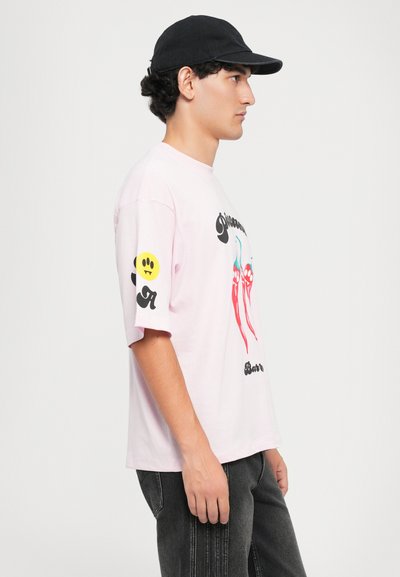 Svetloružové tričko s grafickou potlačou medúzy, čierne písmená a žltý úsmev na ľavom rukáve; nosené s čiernou šiltovkou a šedými nohavicami.