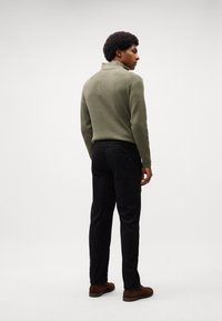 Oliven ribbet høyhals genser, svarte slim-fit bukser, brune sko; modell vender bort, viser frem teksturerte materialer og tettsittende design.
